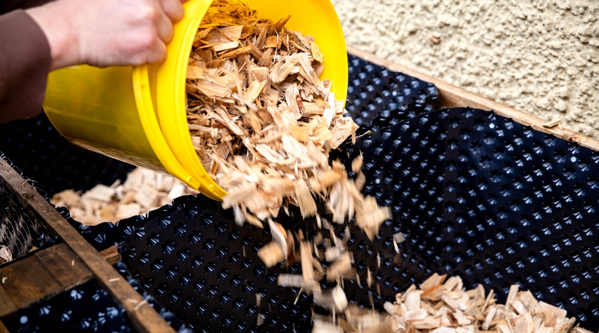 Close-up of spreading mulch (wood chips) into the bottom of a raised bed to prevent dirt and weeds from growing. Wood chips are chopped light wood into small squares.
