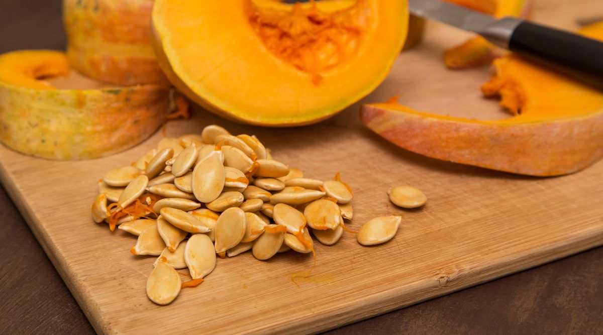 A shot of a sliced pumpkin with its seeds extracted on a wooden cutting board. The pumpkin is in sections in the background, with a kitchen knife sitting on top. The seeds sit in the foreground, unwashed with pumpkin flesh still attached. 