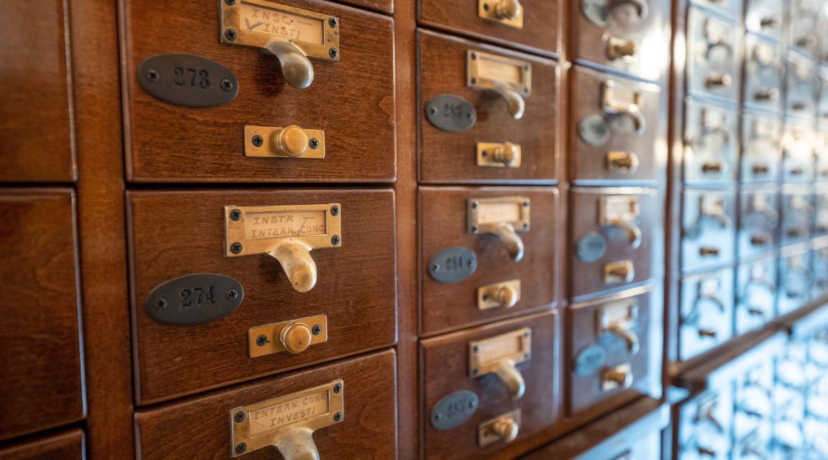 An up-close and offset view of an old wooden card catalog. The handles on each drawer are gold or brassy, and the labels are metal as well, with a deep patina. There are several cabinets stacked on top of one another. 