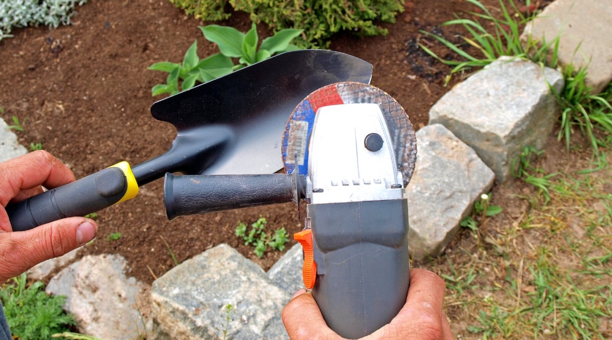 Close-up of a man's hands sharpening a garden trowel with an Angle Grinder. The trowel is a small hand-held gardening tool with a durable black and yellow rubber attached to a sharp black metal blade. The blade is flat, bucket-shaped, resembling a miniature shovel.