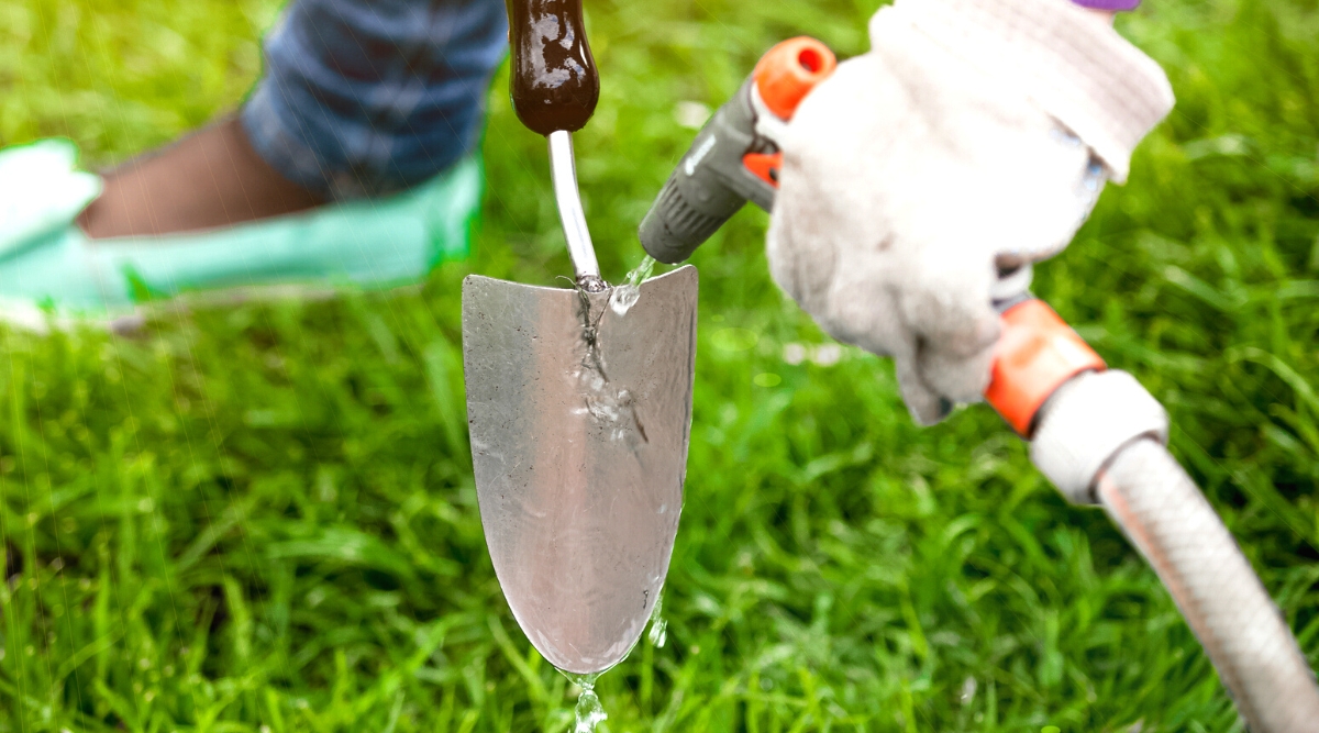 Close-up of a gardener's hands in white gloves washing a garden tool (trowel) with a hose in the garden, against the backdrop of green grass. The gardener is dressed in blue jeans and mint shoes.